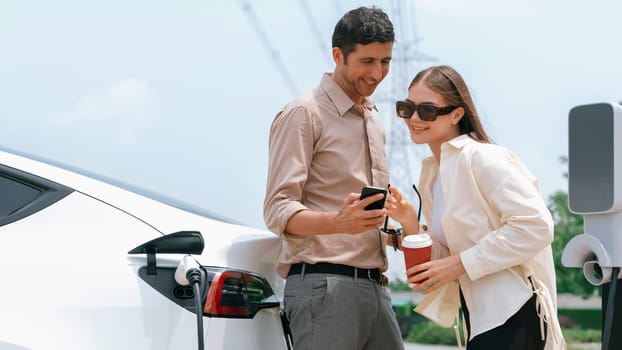 Young couple with coffee pay for electricity while recharge EV car battery at charging station connected to electrical power grid tower as electric industrial for eco friendly car travel. Expedient