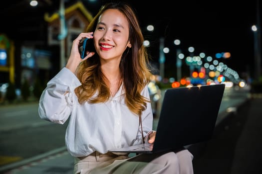 City streets come alive at night, a woman is multitasking on a street corner with her laptop and cell phone, utilizing the power and convenience of modern technology to stay connected with her online