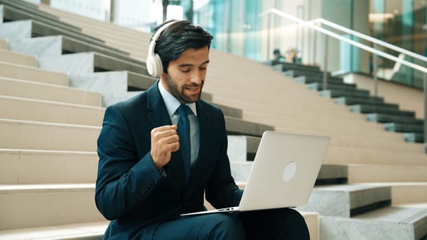 Confident smart business man celebrate successful business idea while listen music. Happy manager project working by using laptop while wearing headphone and suit. Investor sitting at stair. Exultant.