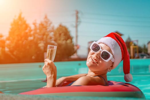 A happy woman in a blue bikini, a red and white Santa hat and sunglasses poses in the pool in an inflatable circle with a watermelon pattern, holding a glass of champagne in her hands. Christmas holidays concept