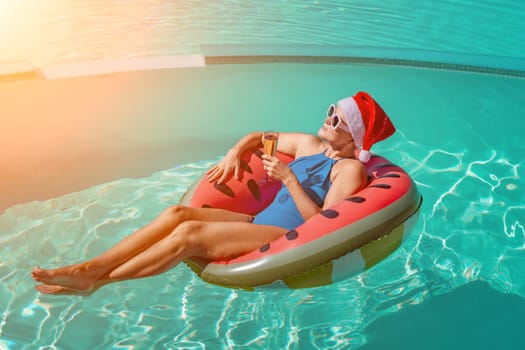 A happy woman in a blue bikini, a red and white Santa hat and sunglasses poses in the pool in an inflatable circle with a watermelon pattern, holding a glass of champagne in her hands. Christmas holidays concept