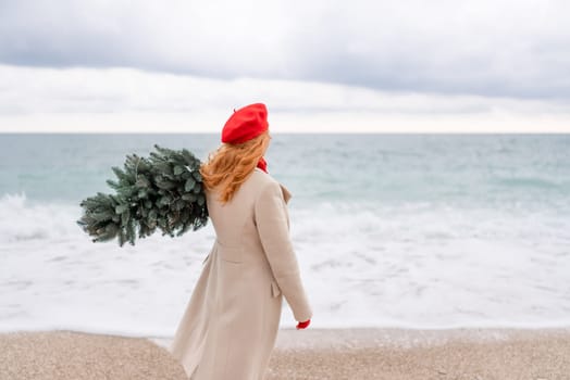 Redhead woman Christmas tree sea. Christmas portrait of a happy redhead woman walking along the beach and holding a Christmas tree on her shoulder. She is dressed in a light coat and a red beret