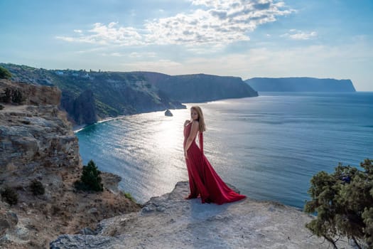 A girl with flowing hair in a long red dress stands on a rock above the sea. The stone can be seen in the sea. Sunny path to the sea from the sun