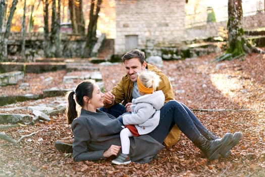 Smiling dad stretches a twig while sitting next to mom with a little girl on her stomach on fallen leaves in the forest. High quality photo