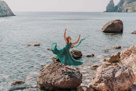 Woman green dress sea. Female dancer in a long mint dress posing on a beach with rocks on sunny day. Girl on the nature on blue sky background