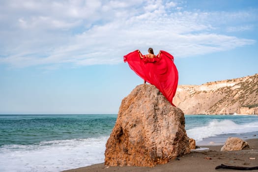 woman sea red dress. Woman with long hair on a sunny seashore in a red flowing dress, back view, silk fabric waving in the wind. Against the backdrop of the blue sky and mountains on the seashore