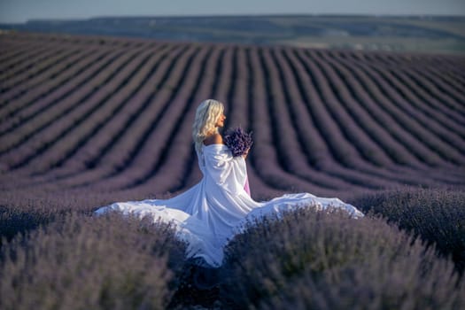 Blonde woman poses in lavender field at sunset. Happy woman in white dress holds lavender bouquet. Aromatherapy concept, lavender oil, photo session in lavender.