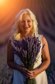 Blonde woman poses in lavender field at sunset. Happy woman in white dress holds lavender bouquet. Aromatherapy concept, lavender oil, photo session in lavender.