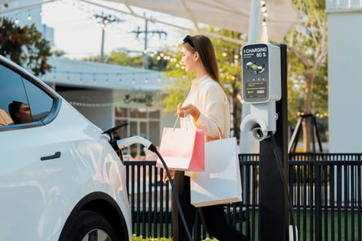 Young woman holding shopping bag recharge EV car battery from charging station at parking lot. Modern woman go shopping by environmental friendly electric vehicle in urban travel lifestyle. Expedient
