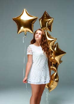 Portrait of a young attractive woman in a white dress, holding bunch of many gold balloons, over light background