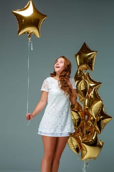 Portrait of a young attractive woman in a white dress, holding bunch of many gold balloons, over light background