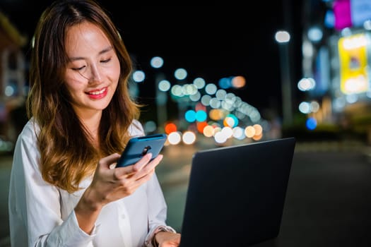 Young Asian woman walks and talks on phone while using laptop computer on city street at night, multitasking with technology.