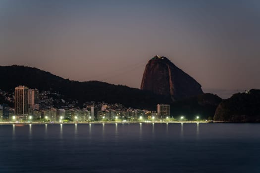 Evening falls on a city, highlighting Sugarloaf Mountain and a lit-up seaside.