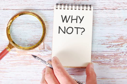 WHY NOT written on a blank sheet of a notebook which is touched by a man's hand with a pencil and a magnifying glass is lying next to it