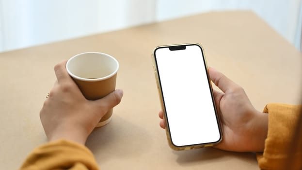 Closeup woman using mobile phone and drinking coffee at desk. Empty screen for application advertising.