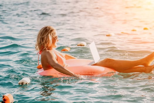 Woman works on laptop in sea. Freelancer, young blond woman in sunglases floating on an inflatable big pink donut with a laptop in the sea at sunset. Freelance, travel and holidays concept