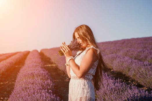 Close up portrait of young beautiful woman in a white dress and a hat is walking in the lavender field and smelling lavender bouquet.