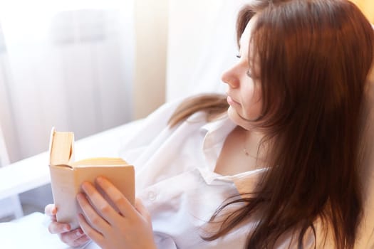 A cute brunette girl is reading a small book of poetry in the bedroom in the morning. A young pregnant woman in the maternity hospital with a book of tips on parenting. A sick and recovering lady