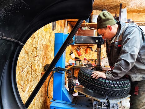 Ishim, Russia - November 27, 2023: Mechanic repairs a car wheel and makes a tire in a dirty garage. Replacement of summer wheels, tires for winter indoors