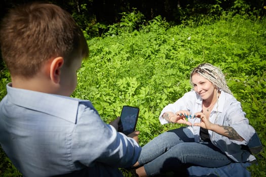 Kirov, Russia - June 12, 2023: Young teenage son takes pictures of his mother on cell phone in nature. Mom and boy having fun in grass in clearing