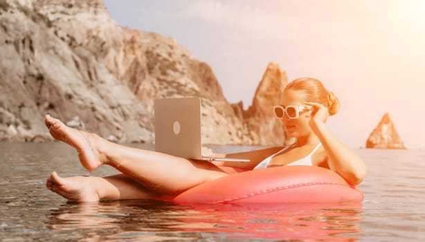 Woman freelancer works on laptop swimming in sea on pink inflatable ring. Pretty lady typing on computer while floating in the sea on inflatable donut at sunset. Freelance, remote work on vacation