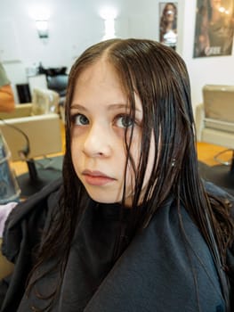 In the bustling atmosphere of a salon, a little girl sits patiently as a stylist with polished red nails carefully trims her damp hair. The sheen of the scissors and the precise hand movements capture the delicate process of styling a child's hair.