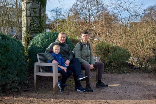 Dad with two sons sitting on a bench in autumn park. Experience the tranquility of familial bonds in the heart of autumn with this serene image. A father, 40 years old, and his two sons - a beautiful 8-year-old boy and a 17-year-old young man, seated in the park. The autumnal ambiance adds warmth to this captivating moment of family togetherness.