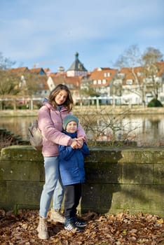 Riverside Family Harmony: Mother, 30 Years Old, and Son - Beautiful 8-Year-Old Boy, Standing by Neckar River and Historic Half-Timbered Town, Bietigheim-Bissingen, Germany, Autumn. Immerse yourself in the picturesque harmony of familial bonds by the riverside with this captivating image. A mother, 30 years old, and her son - a beautiful 8-year-old boy, standing by Neckar River and the historic half-timbered town of Bietigheim-Bissingen, Germany. The autumnal colors add warmth to this timeless family moment.