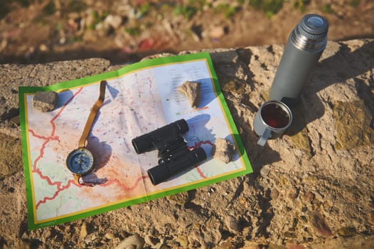 View from above of tourist's clothes or equipment for tourism, a map, compass, binoculars and thermos flask lying down on the rock. Still life