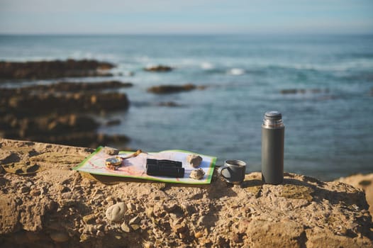 Still life with navigational equipment and thermos flask on the cliff against Atlantic ocean background while waves splashing while breaking on the headland. Compass, binoculars on the tourist's map