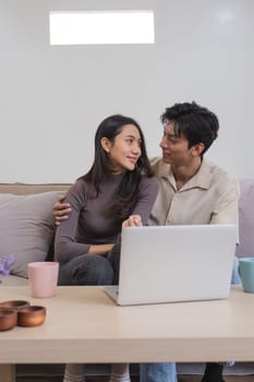 Asian couple watching movies on the internet using laptop at home Smiling Thai man and woman sitting on sofa, hugging each other, looking at computer screen. Surf the web together copy space.