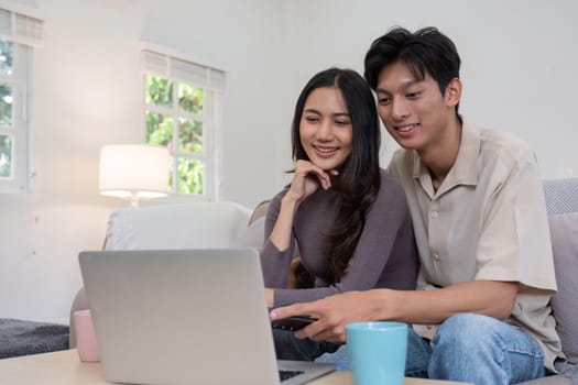 Asian couple watching movies on the internet using laptop at home Smiling Thai man and woman sitting on sofa, hugging each other, looking at computer screen. Surf the web together copy space.