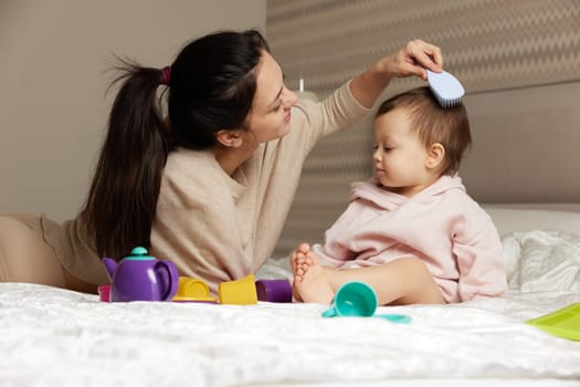 Charming mother is brushing her cute little daughter hair on bed