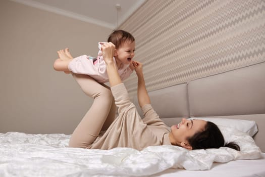 Happy Caucasian mother lifting in air cute little child girl and playing together in bedroom