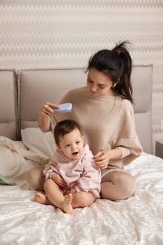 Charming mother is brushing her cute little daughter hair on bed