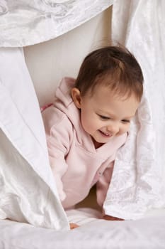 cute funny little child girl looks out from under the blanket on the bed