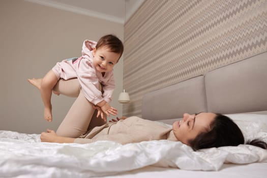 Happy Caucasian mother raising laughing cute baby and playing together in bedroom