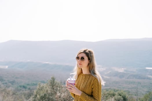 Young woman in sunglasses with a glass of coffee stands high in the mountains and looks into the distance. High quality photo