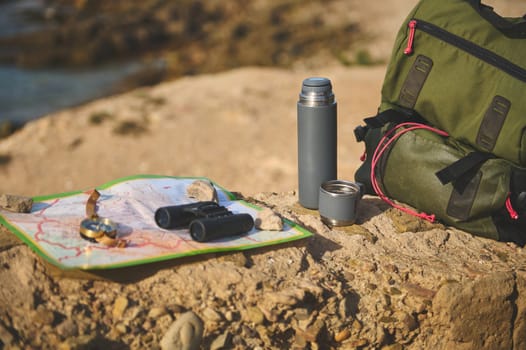 Still life with a green backpack nd thermos flask on the cliff against Atlantic ocean background while waves splashing while breaking on the headland. Compass, binoculars on the tourist's map