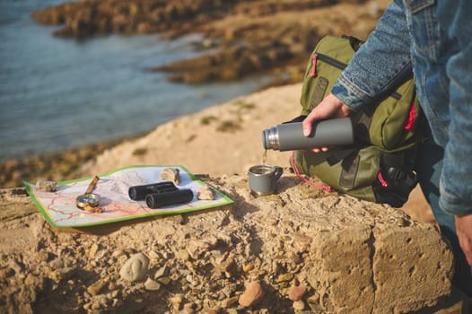 Close-up tourist hand holding thermos flask and pouring hot tea drink into a stainless steel mag, standing on a rock near a map, compass and binoculars. Active lifestyle. Recreation. Tourism concept