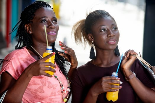 Young girls standing outdoors looking at something up there smiling with shopping bags and bottles of juice in hand.