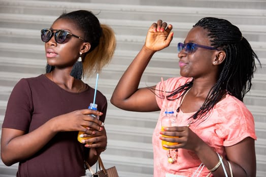 Young girls standing in sunglasses with bottles of fruit juice watching something with shopping bags in hand.