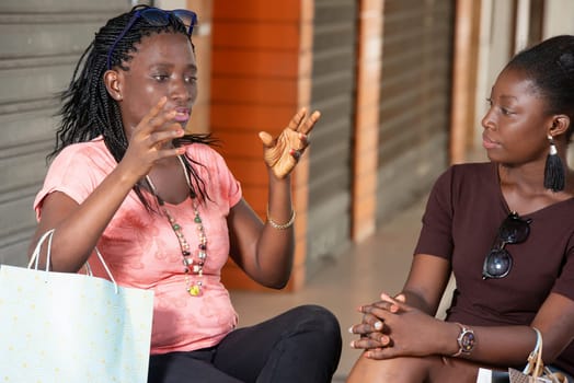 two young women with shopping bags sitting outside are resting after their shopping in an outdoor market, talking, laughing and gesturing.