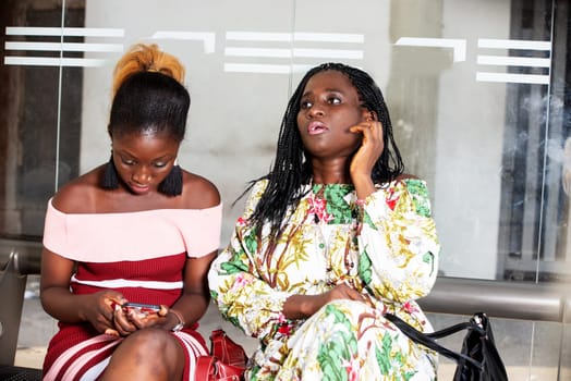 portrait of young woman sitting in a waiting room talking to her girlfriend while this one is watching mobile phone.