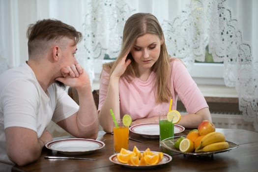 A young girl sits at a table. There are dishes and fruits on the tabletop. Next to the girl sits a young man wearing a white T-shirt.