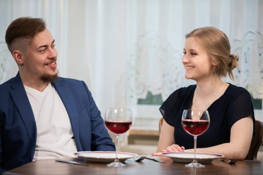 A young married couple is sitting at a table at a family party. The woman smiles at her husband. In the background you can see a window covered with a white curtain.