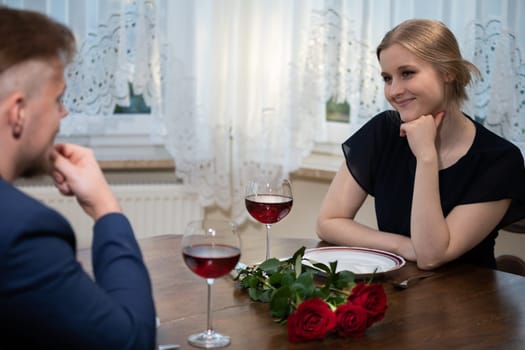 Elegant young girl sits at the table and smiles. She is wearing a black dress. A man wearing a blue jacket sits in front of her. There is a bouquet of red roses on the table.