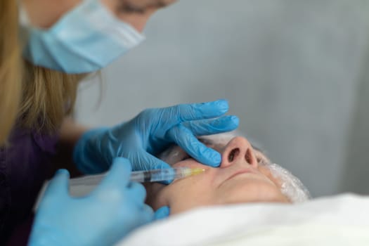 Large close-up of a woman's face seen from below. A woman undergoes a medical procedure. A nurse wearing disposable gloves performs an injection into the patient's face.