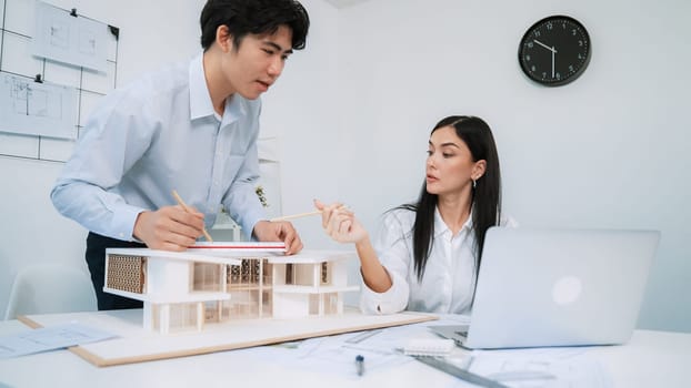 Professional male asian architect using ruler to measure house model length while young beautiful caucasian colleague using laptop to analyzed data on meeting table with house model. Immaculate.