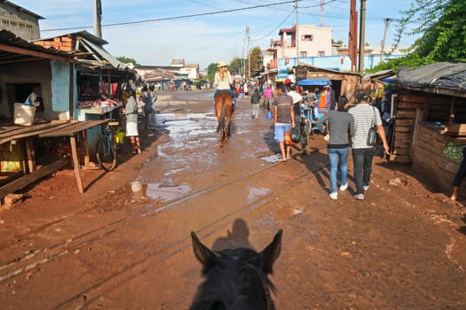 Antsirabe, Madagascar - April 26, 2019: Young woman in straw hat rides on her horse through morning market in Antsirabe, stalls on both sides, car coming in opposite direction, view from her back